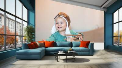 A handsome little boy of 2 years old in a pilot's hat is playing with a wooden airplane and a logistics constructor. Children's wooden toys for child development. Wall mural