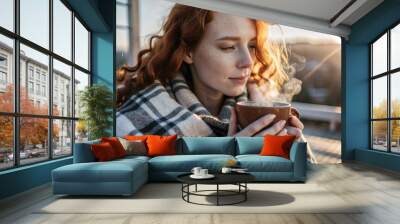 A red-haired, beautiful woman wrapped in a plaid with a mug of hot coffee on a beautiful patio in the rays of the setting sun. The girl inhales the aroma of the drink. Comfort and harmony. Wall mural