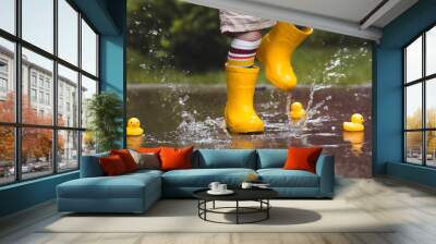 A small child in rainbow-colored socks and yellow rubber boots jumps through puddles and plays with yellow rubber ducks. Legs close-up. A picture of summer and autumn holidays. A child in the rain. Wall mural