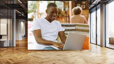 High angle view concentrated multiracial man with laptop sitting on bench with legs in street coffee shop. Editing contracts, documents. Preparing to online webinar, e-learning, doing business course. Wall mural