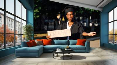 Portrait of poor afro american guy holding cardboard poster and lending hand on street. Looking for job, hiring and employment issue. University or college graduating student in graduate gown and cap. Wall mural