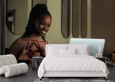 Portrait of young smiling African-American woman with long dark braids wearing pink roll-neck, sitting at table in cafe near laptop, showing heart with fingers, love sign at screen. Communication.  Wall mural