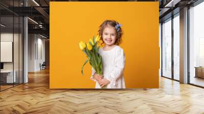 Smiling girl in white dress on yellow background. Cheerful happy child with tulips flower bouquet Wall mural