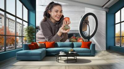 A young woman smiling in the kitchen with a ring lamp and a phone shares healthy food recipes. The concept of Food blogger. Wall mural