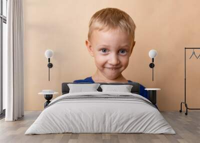 A cheerful young boy with short blonde hair smiling playfully against a simple beige background in a casual blue t-shirt Wall mural