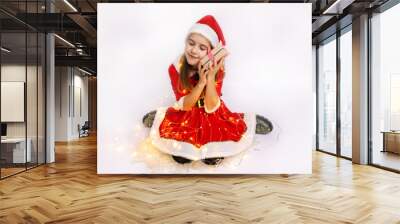 A joyful girl in a festive red dress and Santa hat embraces a gift surrounded by twinkling lights during the holiday season Wall mural