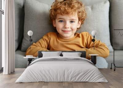 A young boy with curly hair sitting comfortably on a couch, smiling warmly in a cozy living room during the afternoon Wall mural