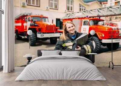 Female firefighter in in a protective suit with documents standing ain the background of a fire engines. Fire station Wall mural