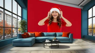 Funny young Santa woman wearing red dress Christmas hat listening music with headphones keeping eyes closed isolated on red background studio portrait. Happy New Year celebration merry holiday concept Wall mural