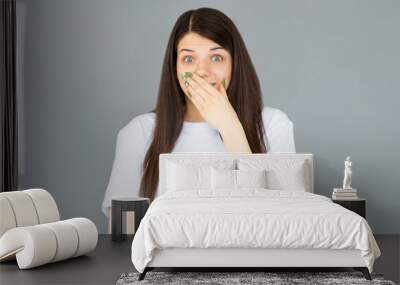 Portrait of a lovely cheerful young girl wearing white t-shirt standing isolated over gray background, covering mouth Wall mural