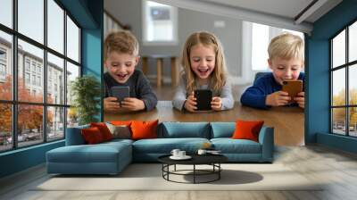 Three children joyfully using smartphones while sitting at a table, showcasing modern technology and playful interaction. Wall mural