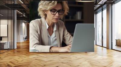 A thoughtful middle-aged businesswoman jotting down notes in a journal while reviewing her laptop screen, her face etched with determination Wall mural