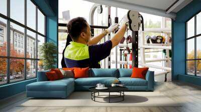 Professional Asian male service worker or fitter checks equipment, maintains and secures fitness equipment in indoor gym, provides safety for users : Skilled technicians repair exercise machines. Wall mural