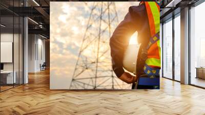 An electrician at a power station wearing a safety suit and hard hat prepares to inspect and maintain electrical systems at high voltage pylons. Wall mural