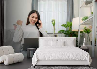 Asian woman working from home With laptop and smart phone sitting on sofa in living room at home and she is excited about shopping online. Wall mural