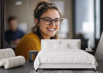 Joyful woman in yellow sweater and glasses, smiling while working on digital marketing project for online business, with blurred office background. Wall mural