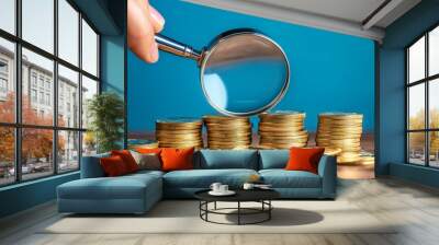 Magnifying glass held over a stack of gold coins on a wooden table with a blue background. This high-resolution image captures superb detail and hyper-realism, ideal for financial themes and marketing Wall mural