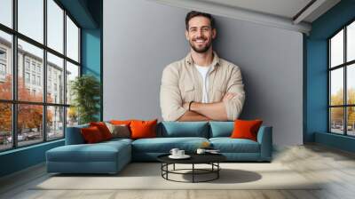 Portrait of a smiling young man in a beige shirt, standing with arms crossed, looking casually at the camera against a gray background. Wall mural