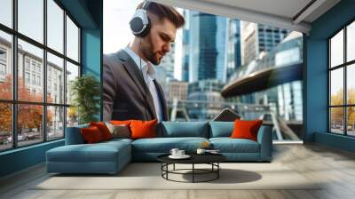 Young businessman in a suit using a tablet and headphones with a modern skyscraper and cityscape background, sharp focus, and bokeh effect. Wall mural