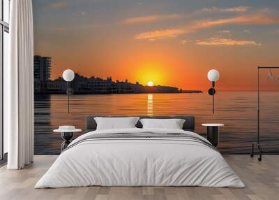 Sunset over the horizon illuminating buildings beside tranquil, reflective coastal waters Wall mural