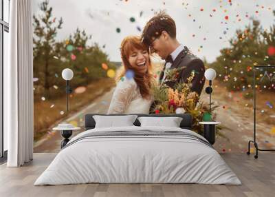 A happy wedding couple is standing on the side of an empty country road, throwing colorful confetti into their air as they smile and embrace each other in celebration Wall mural